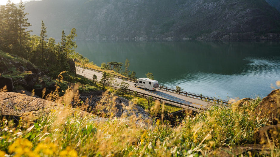 A white motorhome travels along a winding road beside a calm lake, surrounded by steep mountains, trees, and sunlit grasses in the foreground.