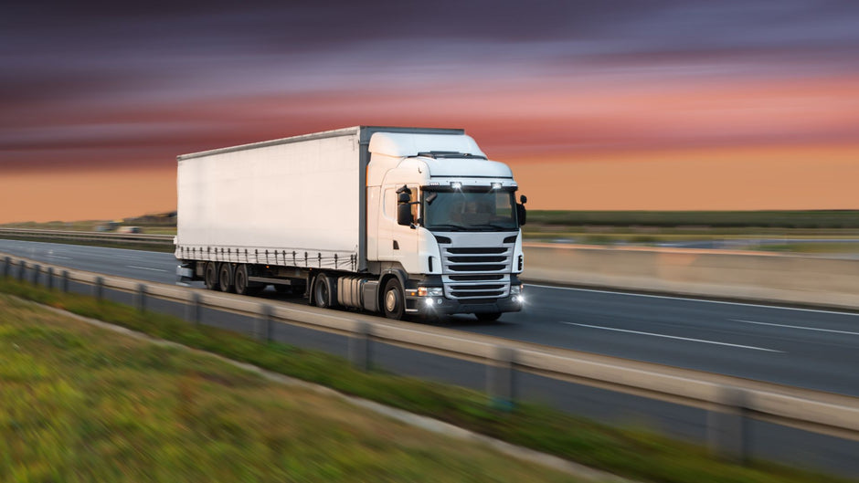 A white semi-truck with a long cargo box driving on a highway during sunset, with blurred grass and road in the background.