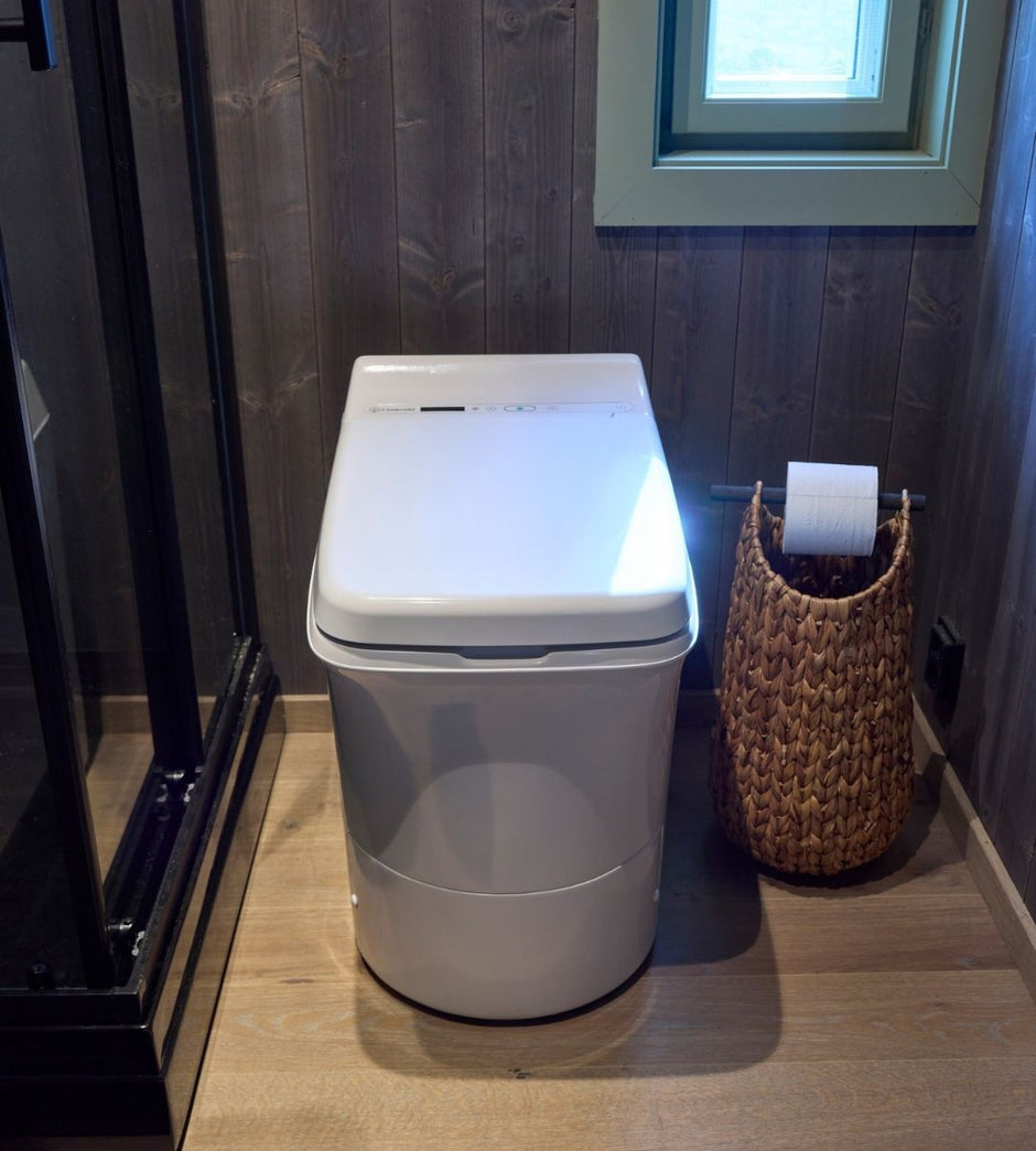 White Cinderella incineration toilet in a bathroom with a woven basket and toilet paper roll.