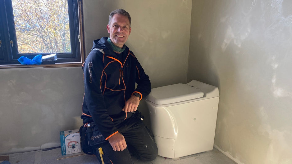 Hombre arrodillado junto a un retrete de incineración de Cinderella en una habitación con paredes inacabadas.