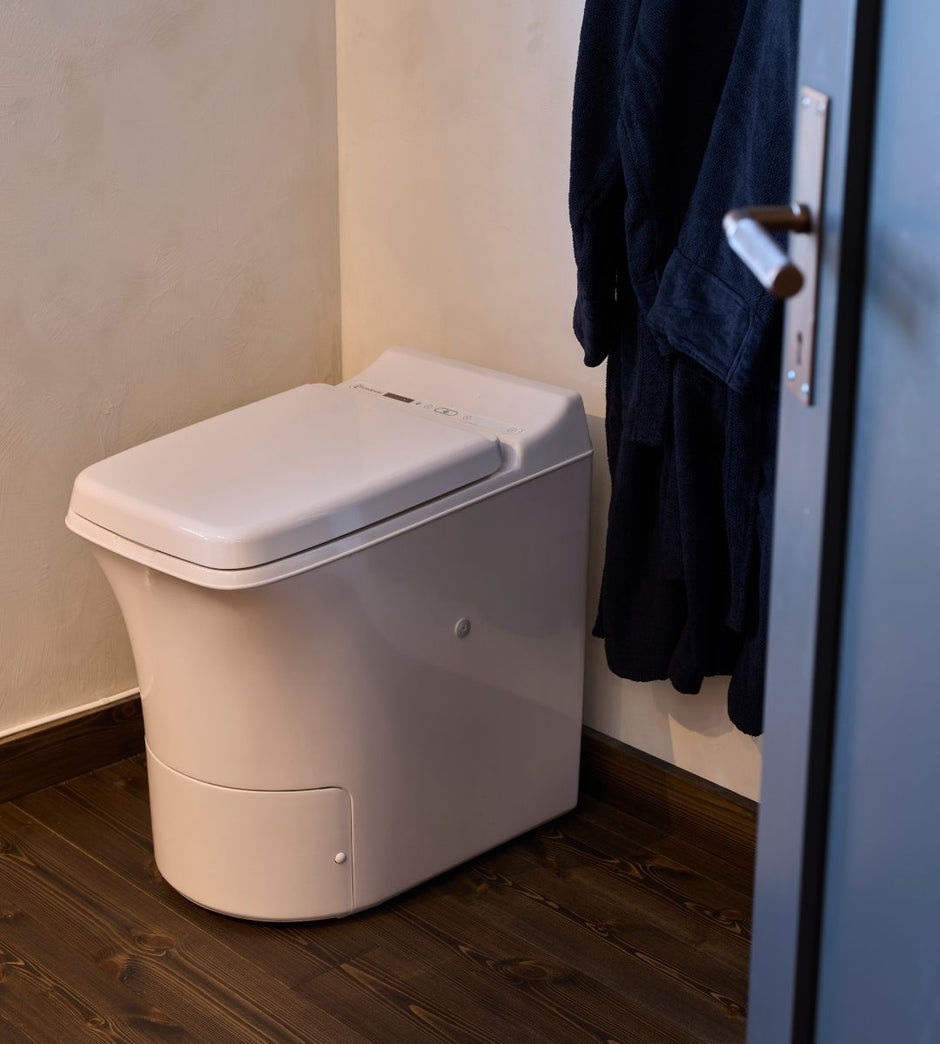 Cinderella Freedom forbrenningstoalett mot en beige vegg med en mørk morgenkåpe hengende på en krok.